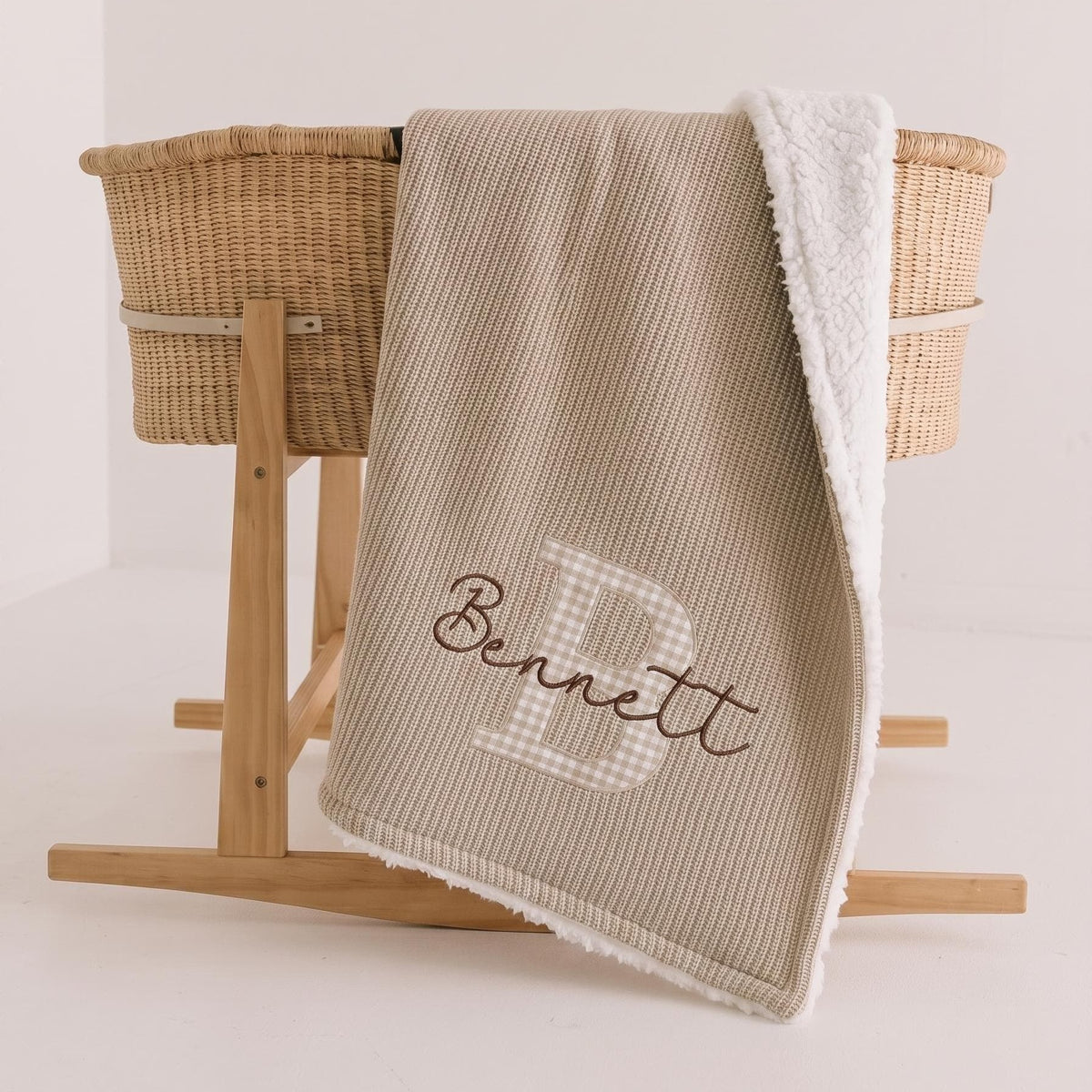 Beige personalized sherpa baby blanket with beige gingham applique letter B and brown embroidered name Bennett draped over a Moses basket