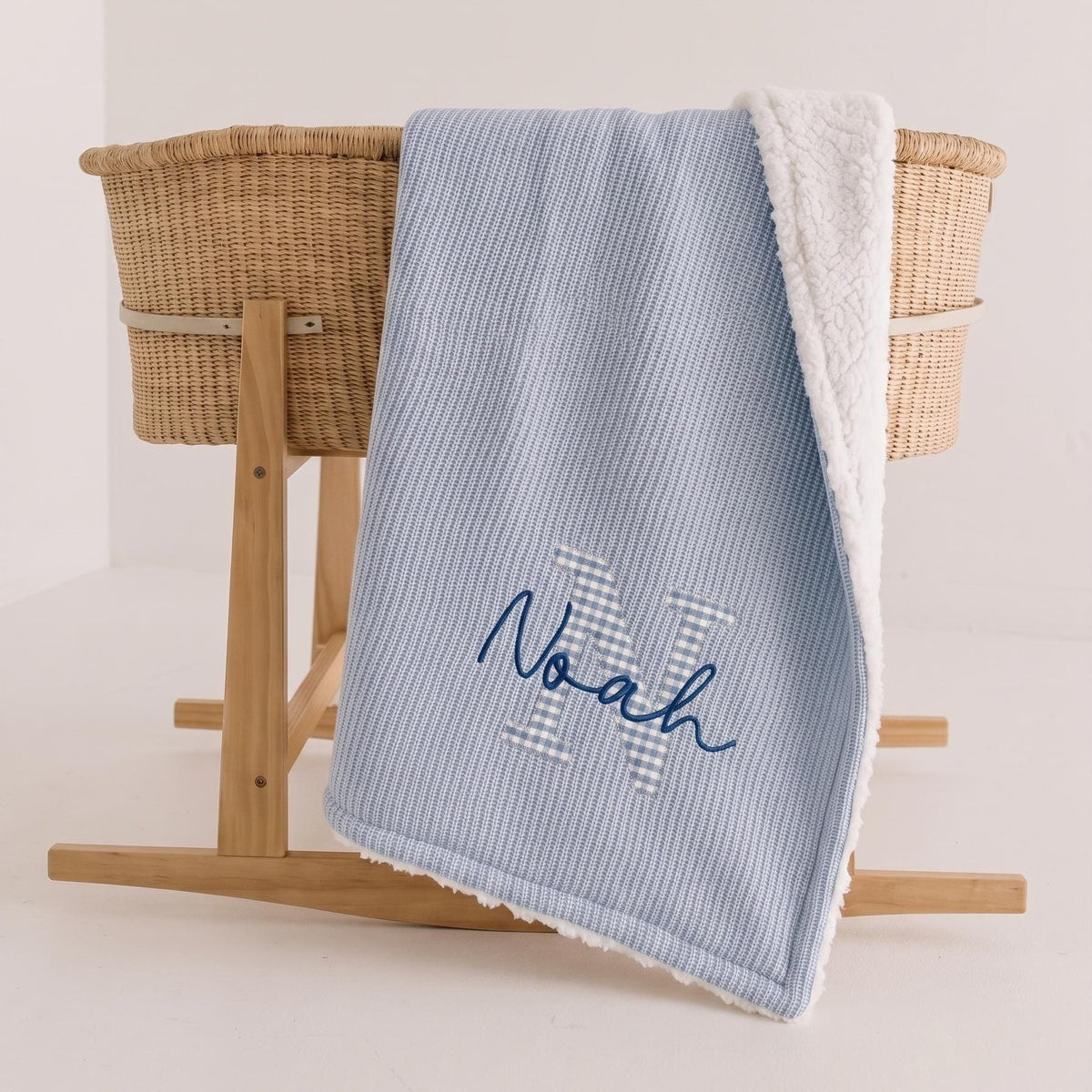 Blue personalized sherpa baby blanket with blue gingham applique letter N and navy embroidered name Noah draped over a Moses basket