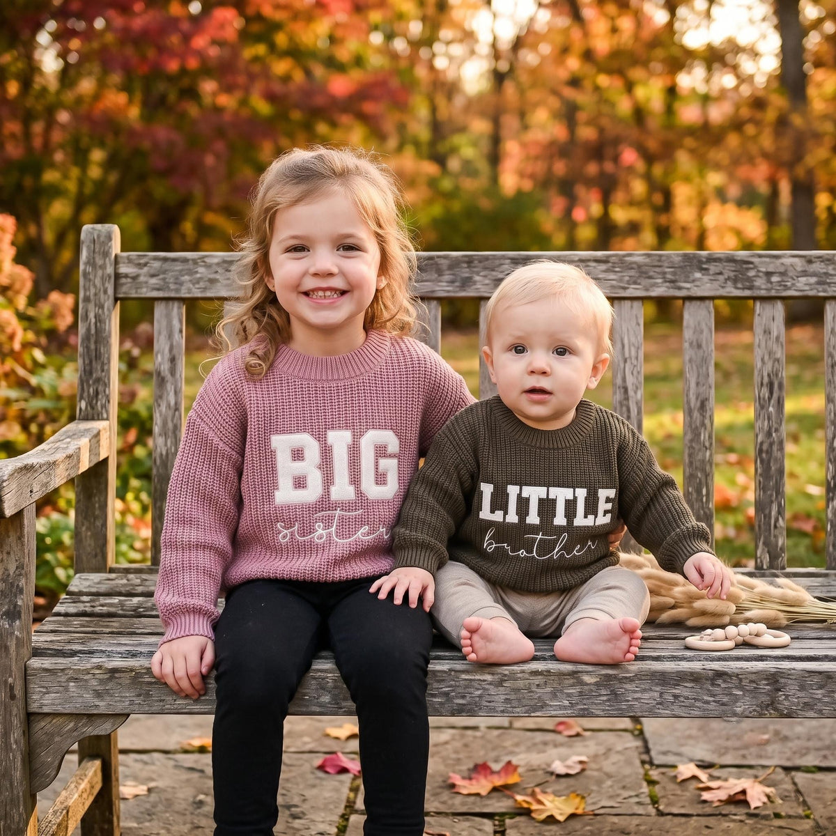 Big sister and little brother in matching sweaters on a bench outdoors in autumn
