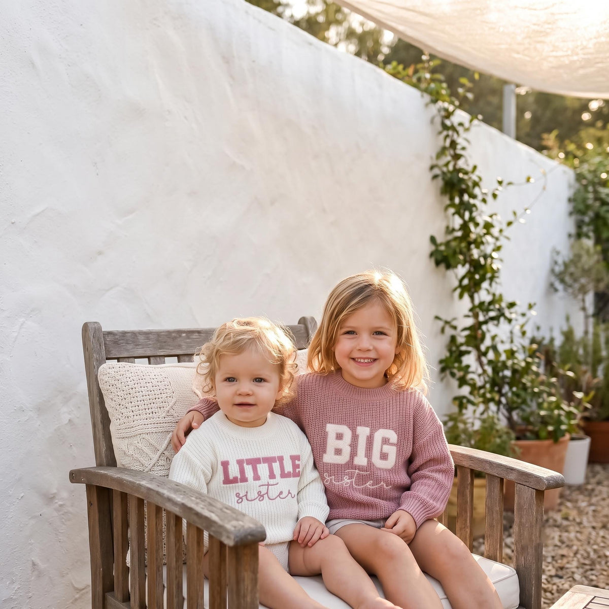 Big sister and little sister in matching embroidered sweaters on a garden bench