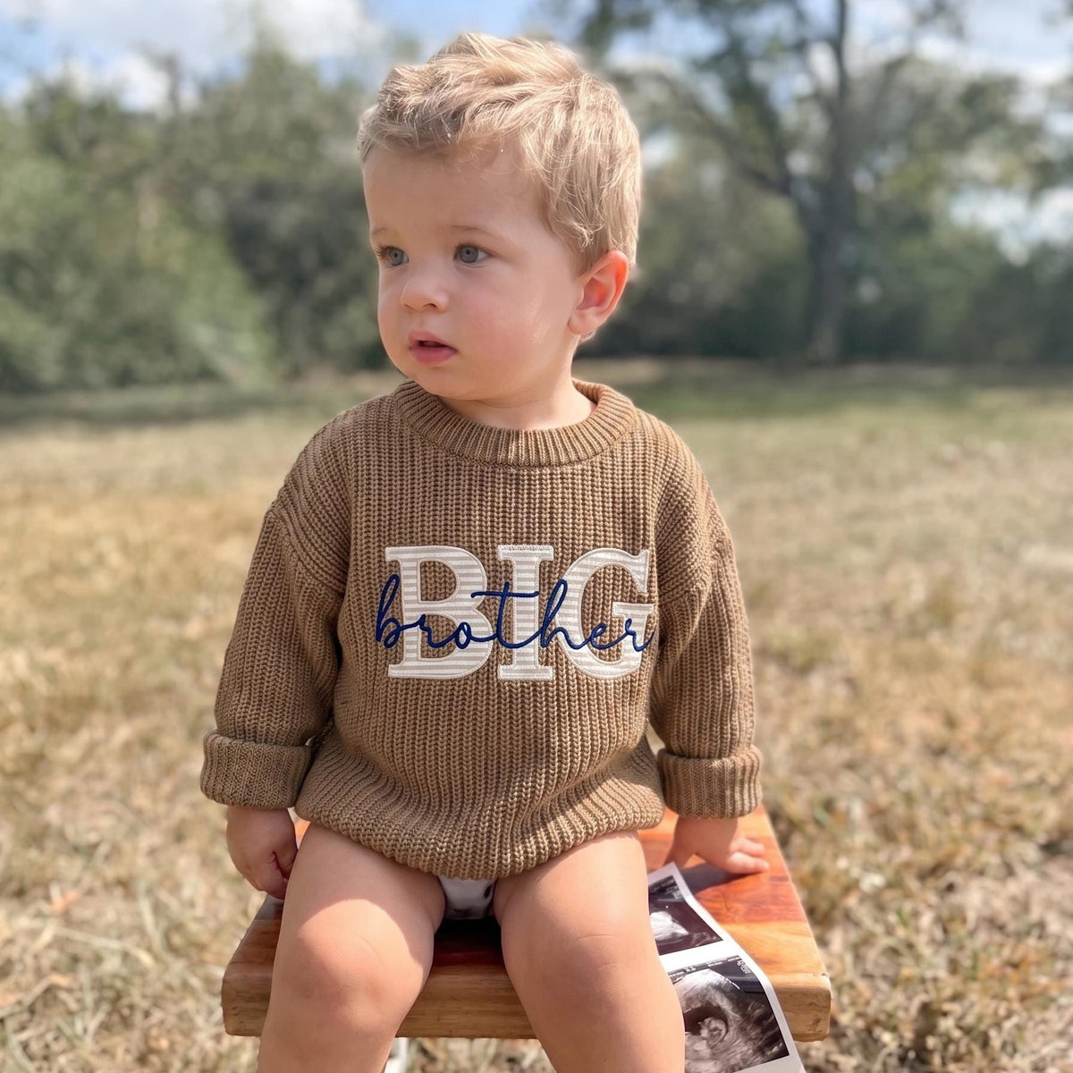 Toddler boy wearing a khaki knit sweater with BIG fabric applique letters and brother embroidered in navy script, sitting outdoors