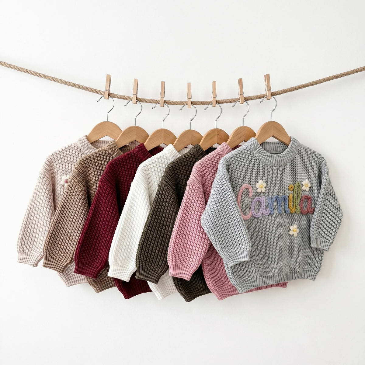 Seven sweater colors hanging on a line: beige, khaki, red, white, olive, pink and sky grey, front sweater embroidered with Camila
