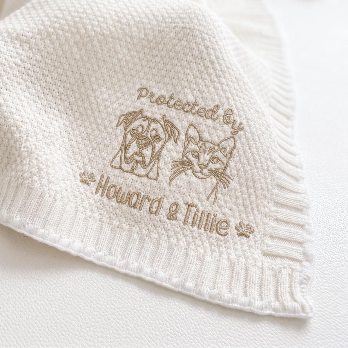 Close-up of embroidered dog and cat line portraits and "Protected by Howard & Tillie" on a knit baby blanket