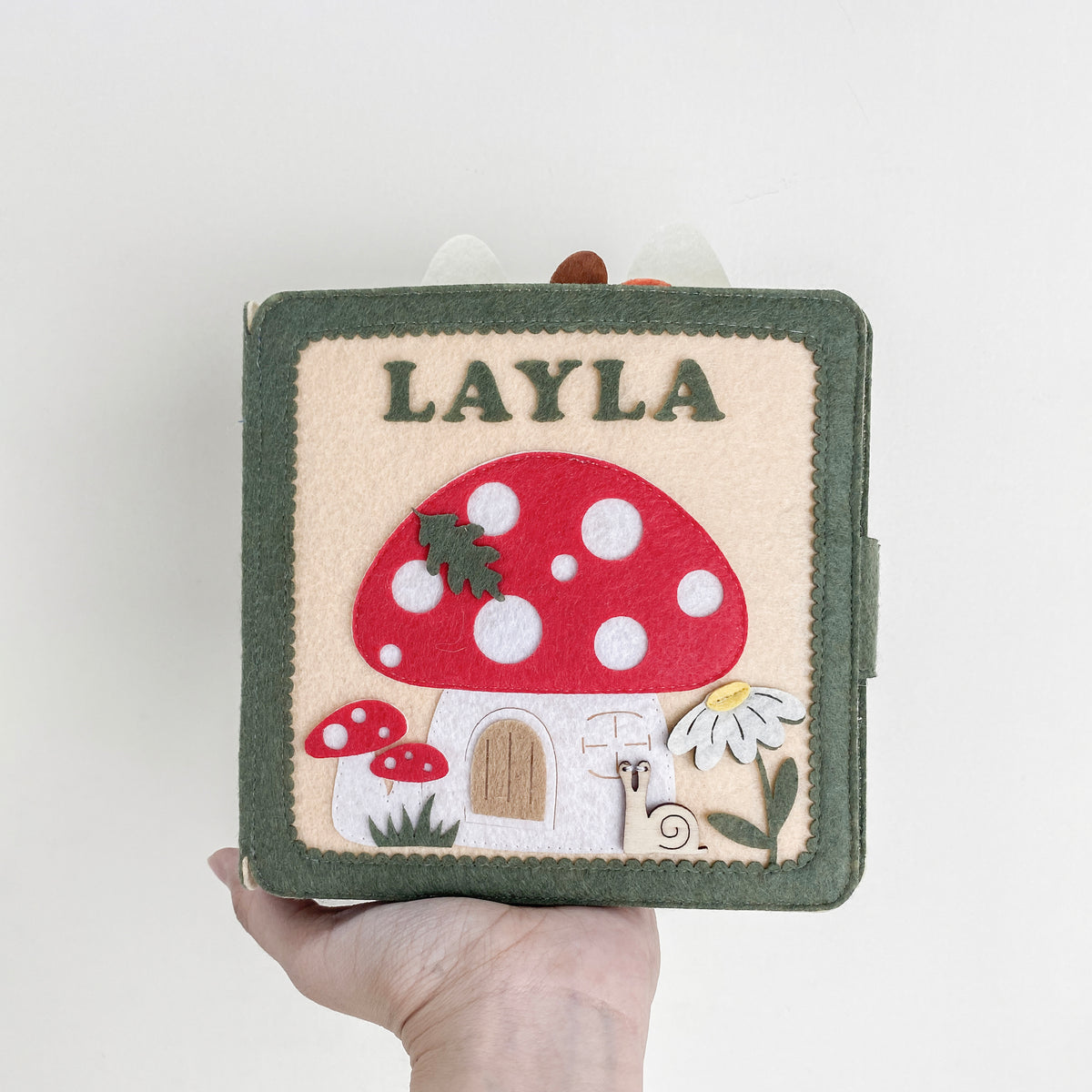 Hand holding a personalized felt quiet book with name Layla and a red mushroom house cover