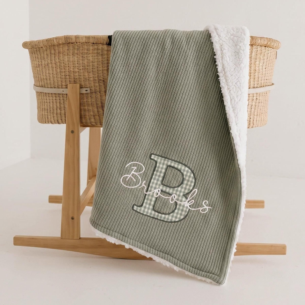 Sage personalized sherpa baby blanket with sage gingham applique letter B and white embroidered name Brooks draped over a Moses basket