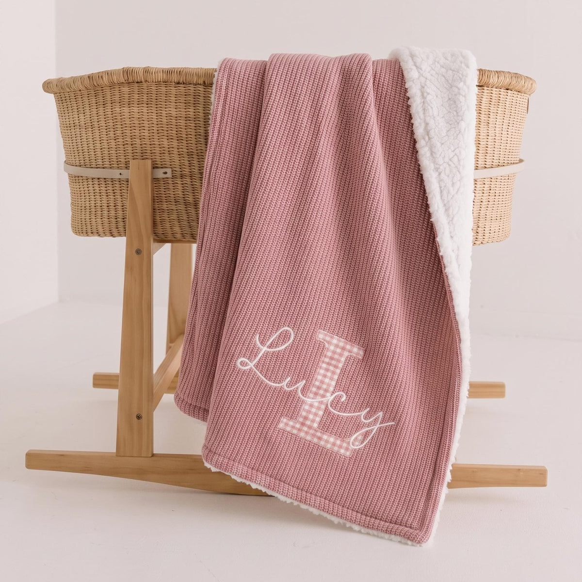 Pink personalized sherpa baby blanket with pink gingham applique letter L and white embroidered name Lucy draped over a Moses basket