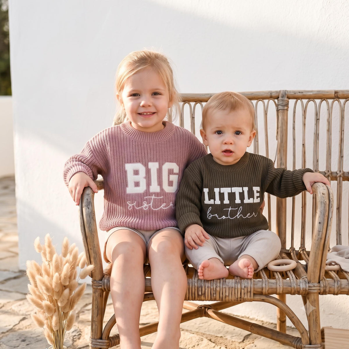 Big sister and little brother in matching embroidered sweaters on a rattan bench