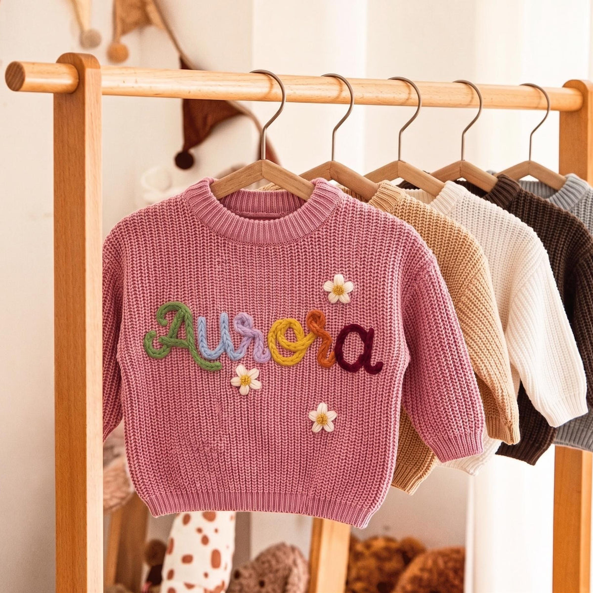 Pink sweater with hand embroidered name Aurora hanging on a wooden clothes rack