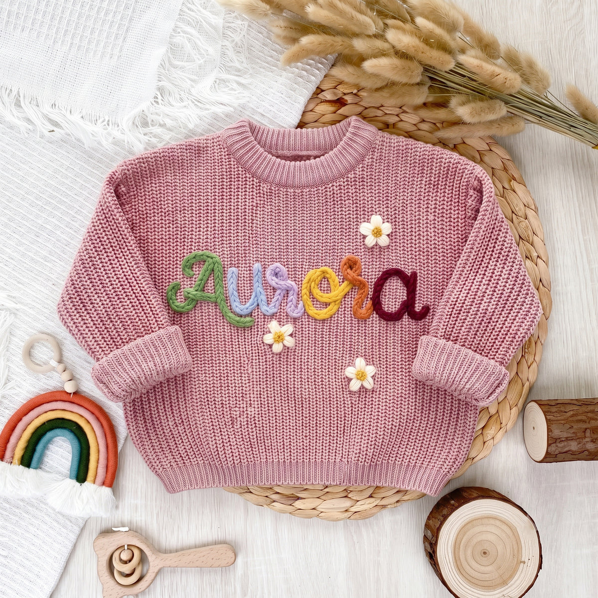 Pink personalized sweater with hand embroidered multicolor yarn name Aurora and flowers, laid flat