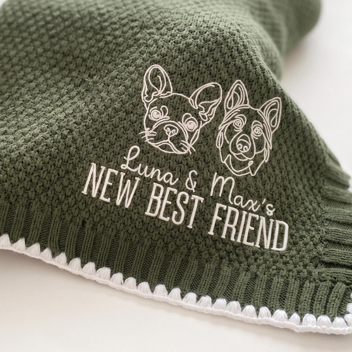 Close-up of embroidered dog and cat line portraits and "Luna & Max's New Best Friend" on an olive knit baby blanket