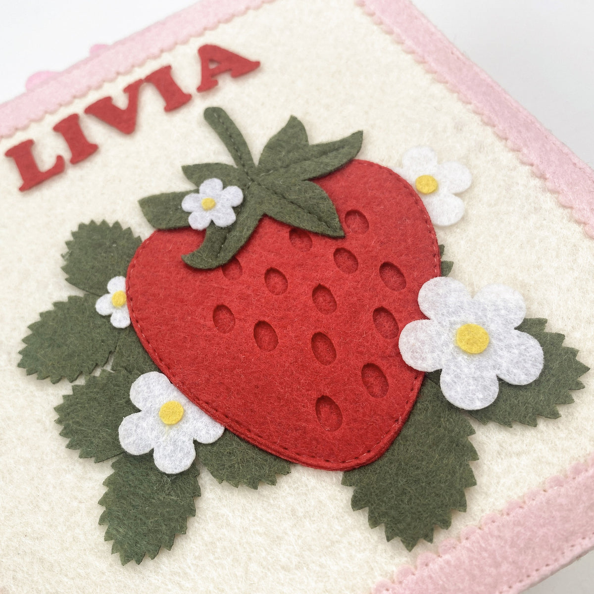 Close-up of felt strawberry cover with embroidered name Livia, white flowers and green leaves